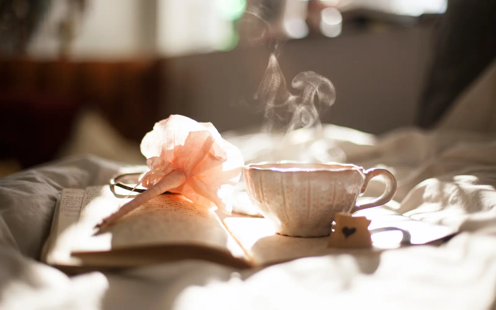 A steaming cup of tea beside a soft flower and an open book on a white bed