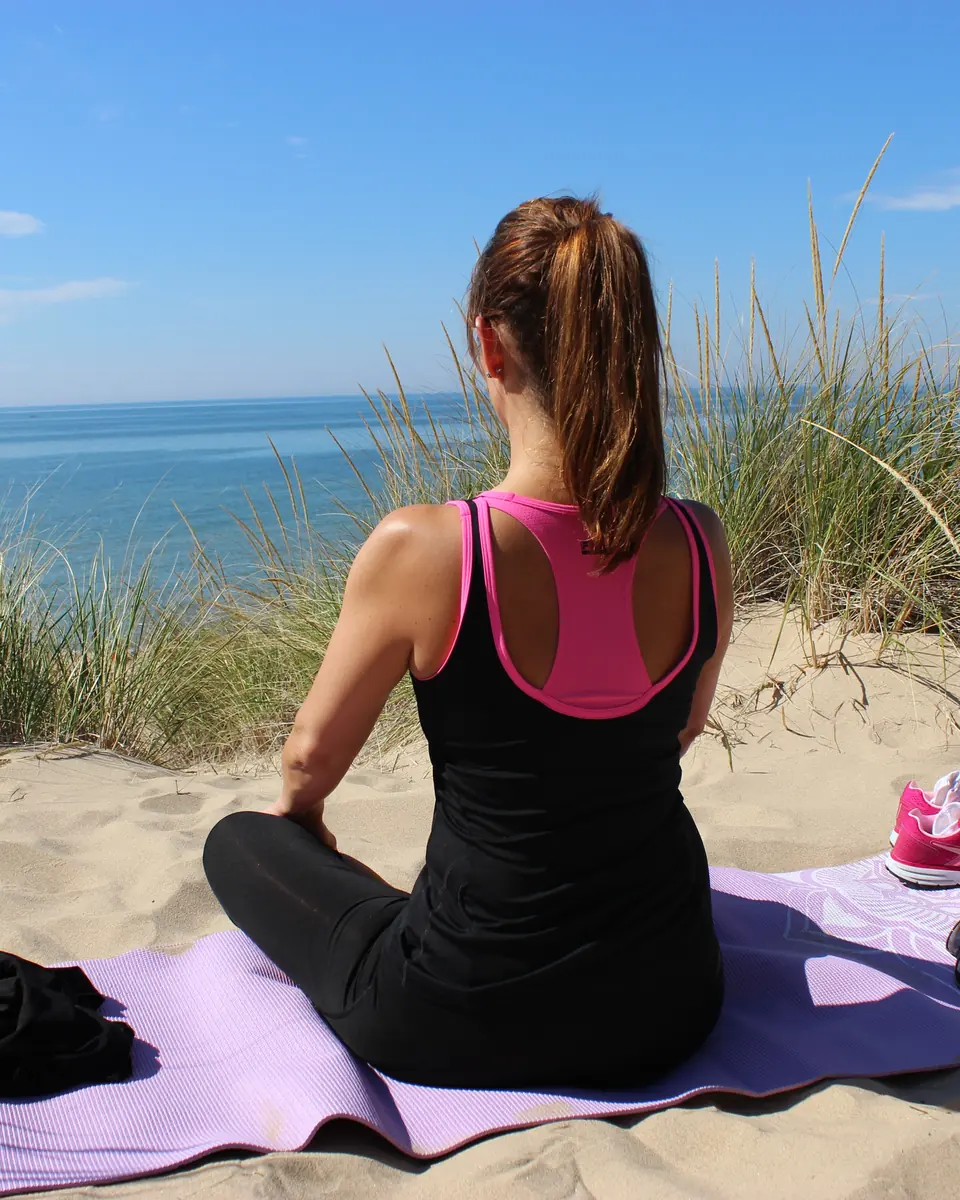 Woman seated cross-legged on a mat in the dunes, facing the sea with her back to the camera