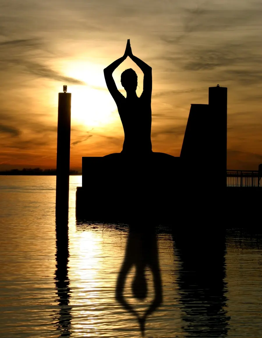 Silhouette meditating on a jetty post at sunset, mirrored in the water