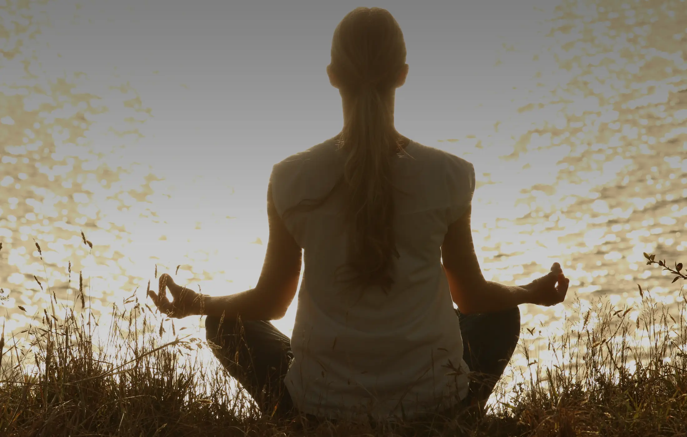 A woman meditating cross-legged in tall grass, backlit by the setting sun